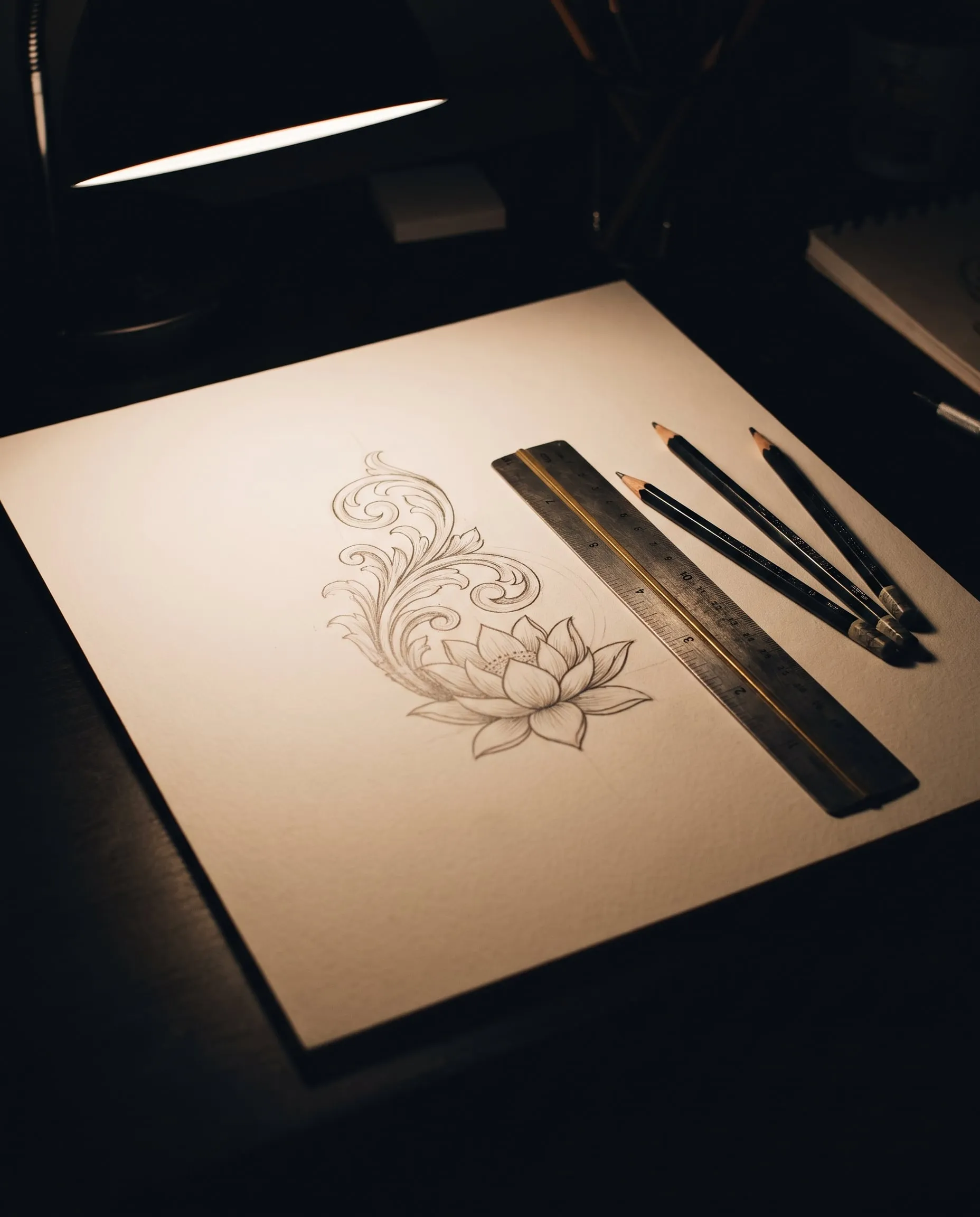 ภาพวาดดินสอรูปดอกบัวและลายกนก ข้างไม้บรรทัดเหล็กและดินสอใต้โคมไฟ